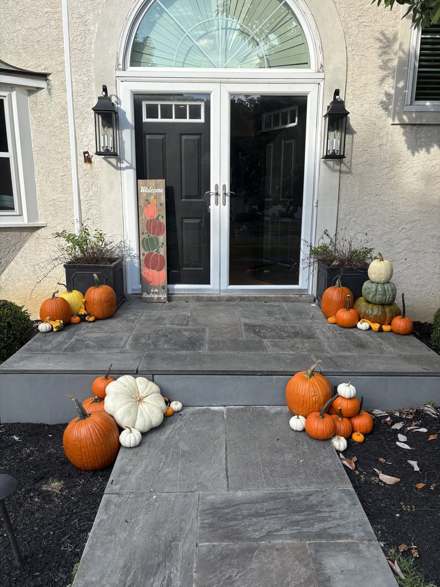 A tall painted welcome sign propped beside black double doors, with pumpkins massed across the flagstone steps and walkway