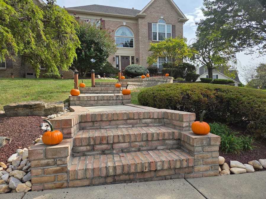 Single pumpkins set along the coping of tiered brick steps and retaining walls climbing to a brick house