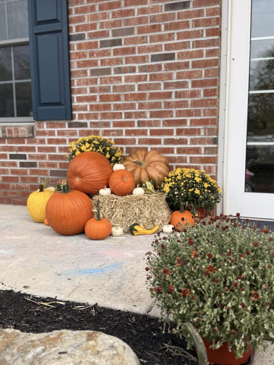 Pumpkins, gourds and yellow mums banked around a straw bale against a brick wall beside a glass door