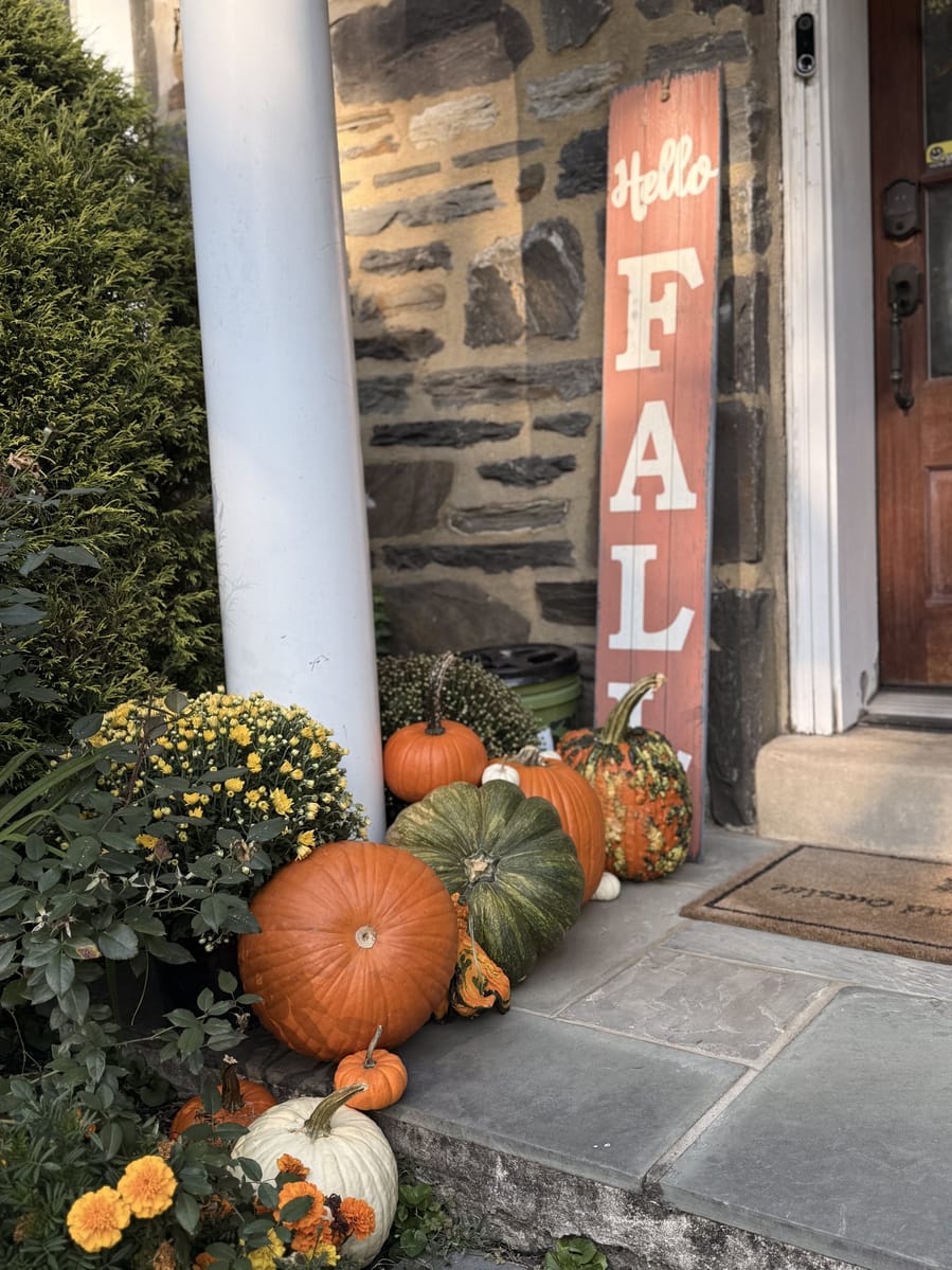 A tall painted Hello Fall sign leaning against stone beside a run of pumpkins, gourds and yellow mums