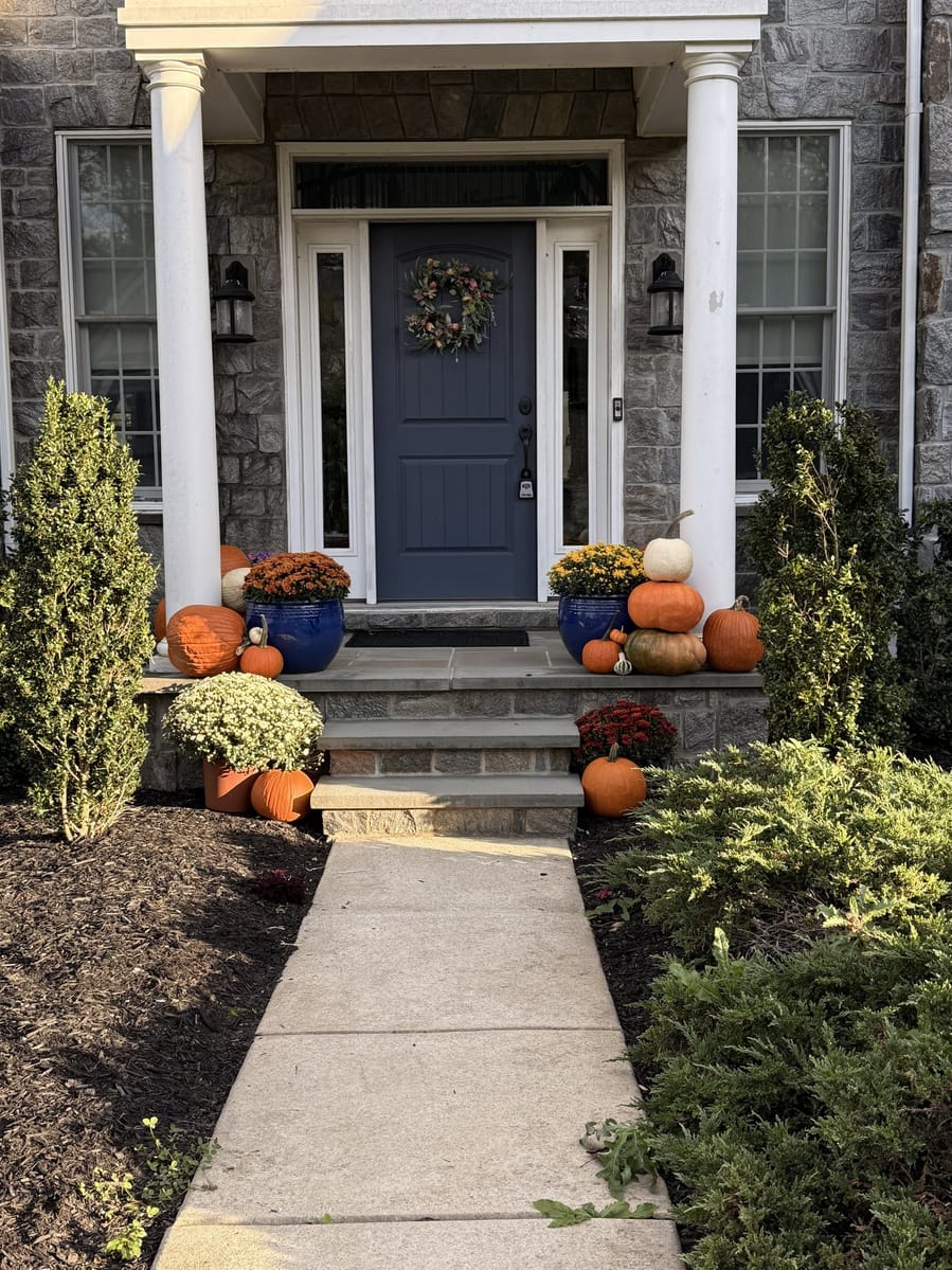 A navy front door with a fall wreath, flanked by blue glazed urns of mums and stacked pumpkins on stone steps