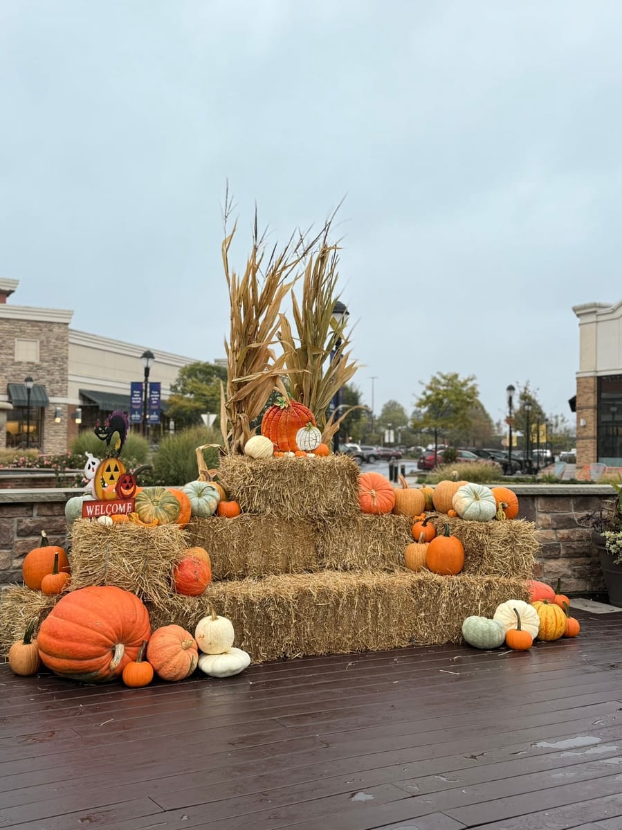 A tiered straw-bale display topped with cornstalks and heirloom pumpkins outside a shopping-center storefront