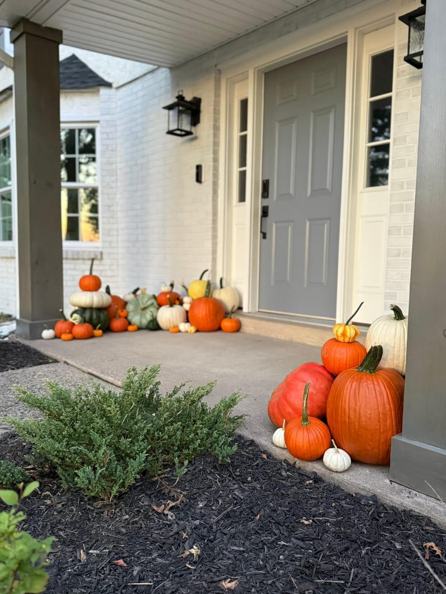 Pumpkins and gourds running the length of a covered porch beside a grey front door, with a second group at the column