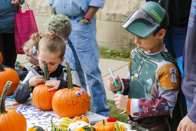 Children in costume decorating pumpkins with markers and googly eyes at an outdoor table