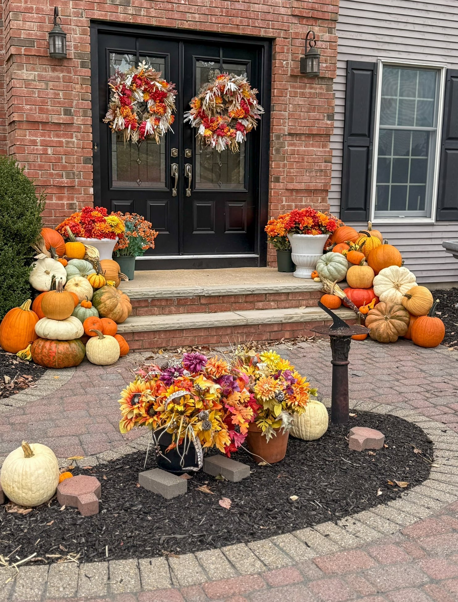 Two banks of pumpkins and gourds flanking the black double doors of a brick home, with fall wreaths on both doors and mums in white urns