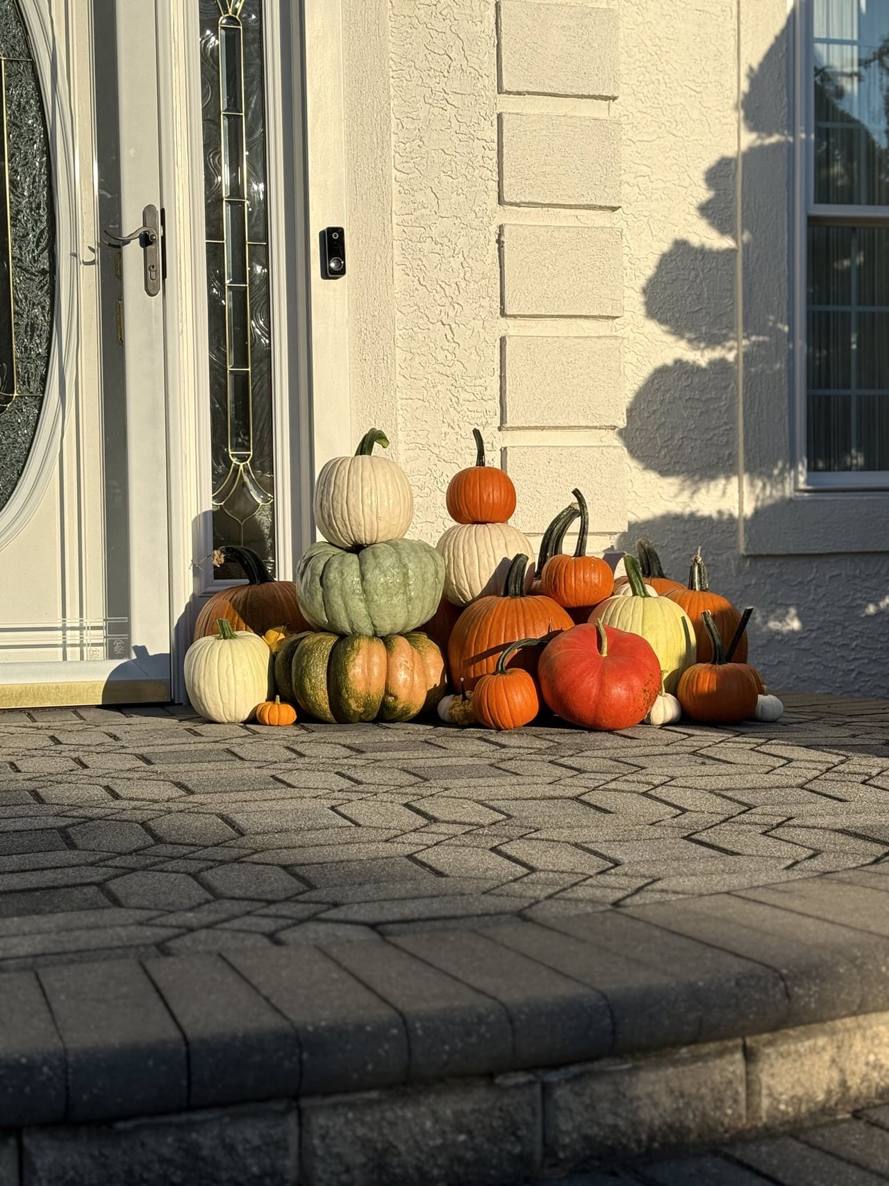 Stacked heirloom and orange pumpkins beside a white front door, lit by low evening sun